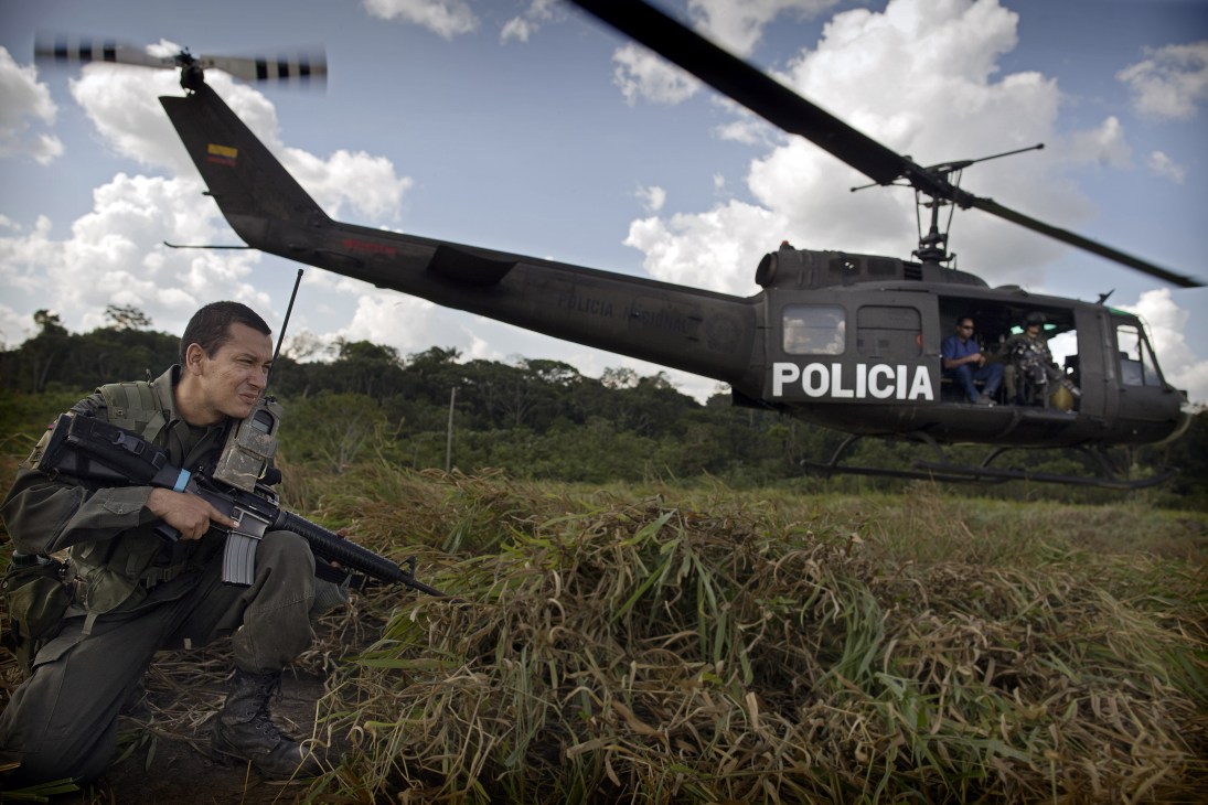 A Colombian police officer crouches with an assault rifle as a police helicopter lands in a jungle clearing.