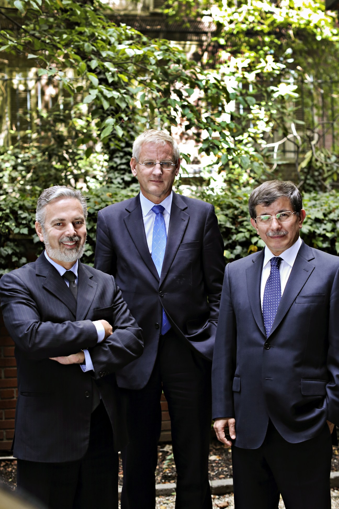 Three foreign ministers in dark suits stand together in a lush garden setting.