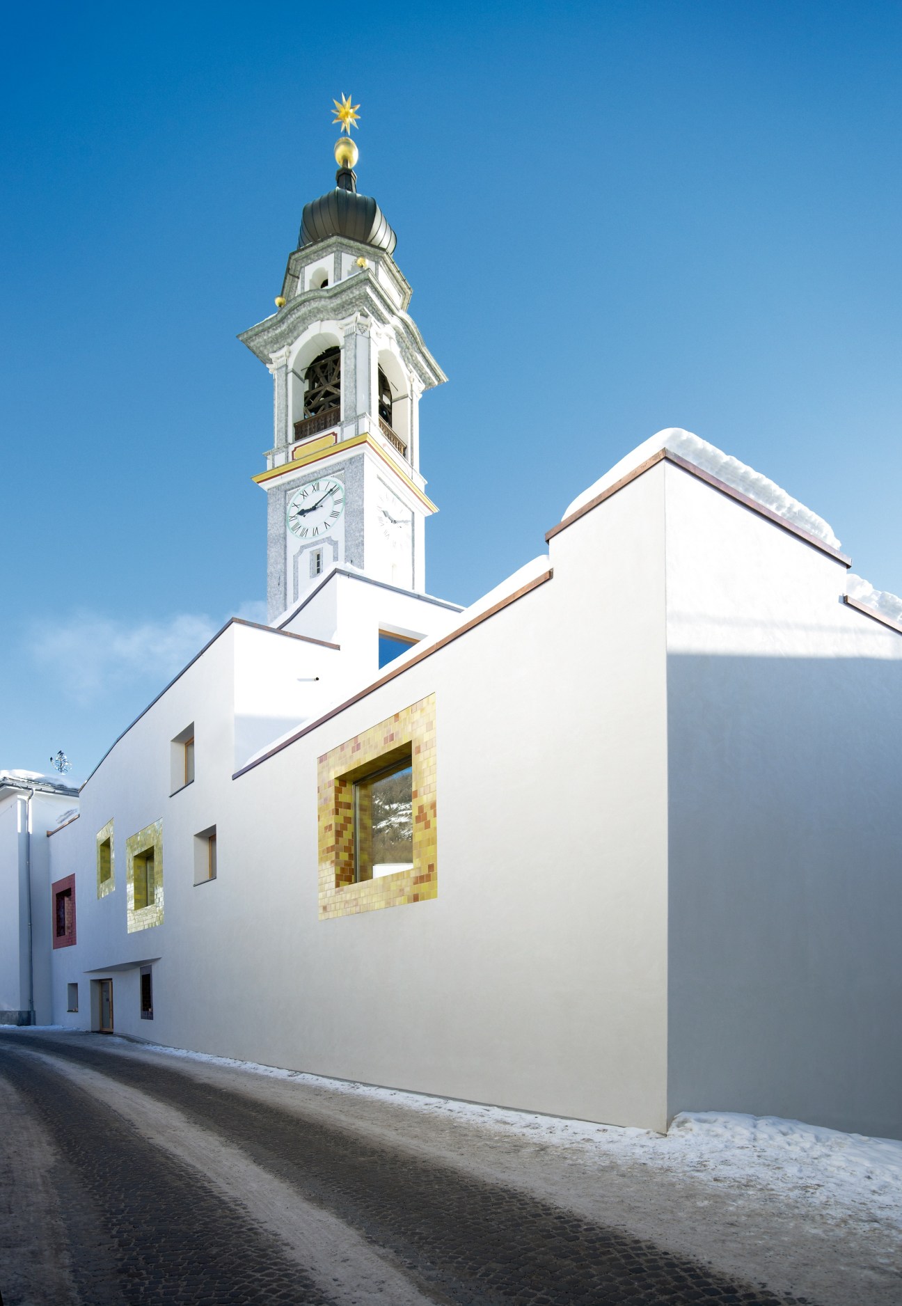 White spa building with golden-framed windows next to a snow-covered church tower in Samedan, Switzerland.