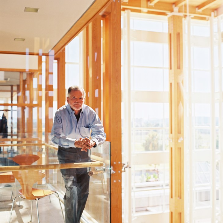 Jim Hudson stands in a modern wooden-framed office with large windows overlooking a landscape.