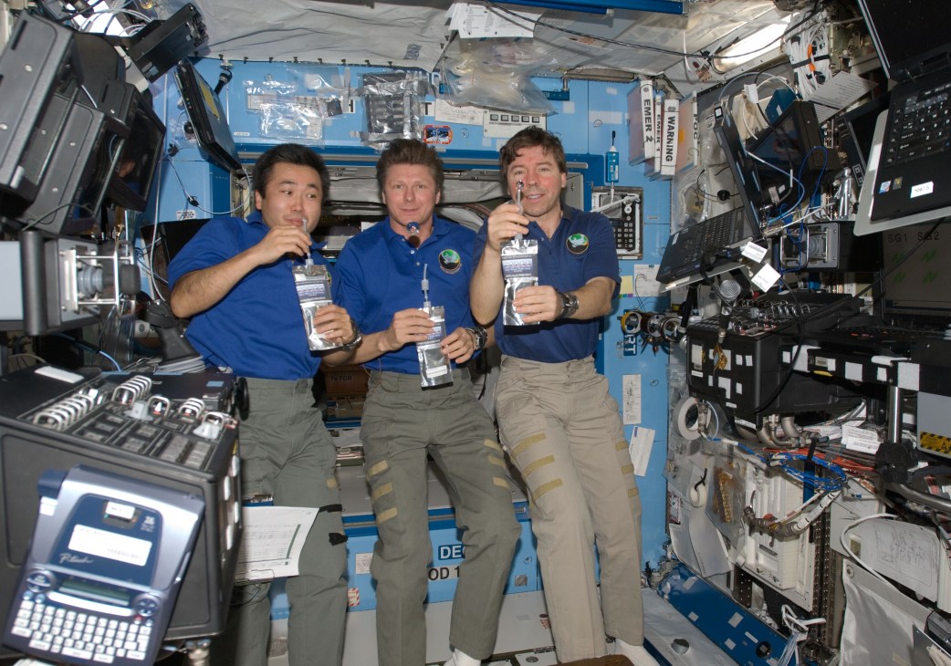 Three astronauts in blue flight suits drinking from pouches aboard the International Space Station.