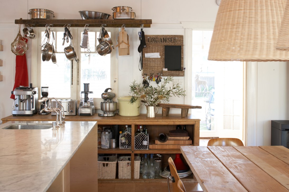 Kitchen with open wooden cabinets, stainless steel cookware hanging above a wooden shelf, and natural light from windows.