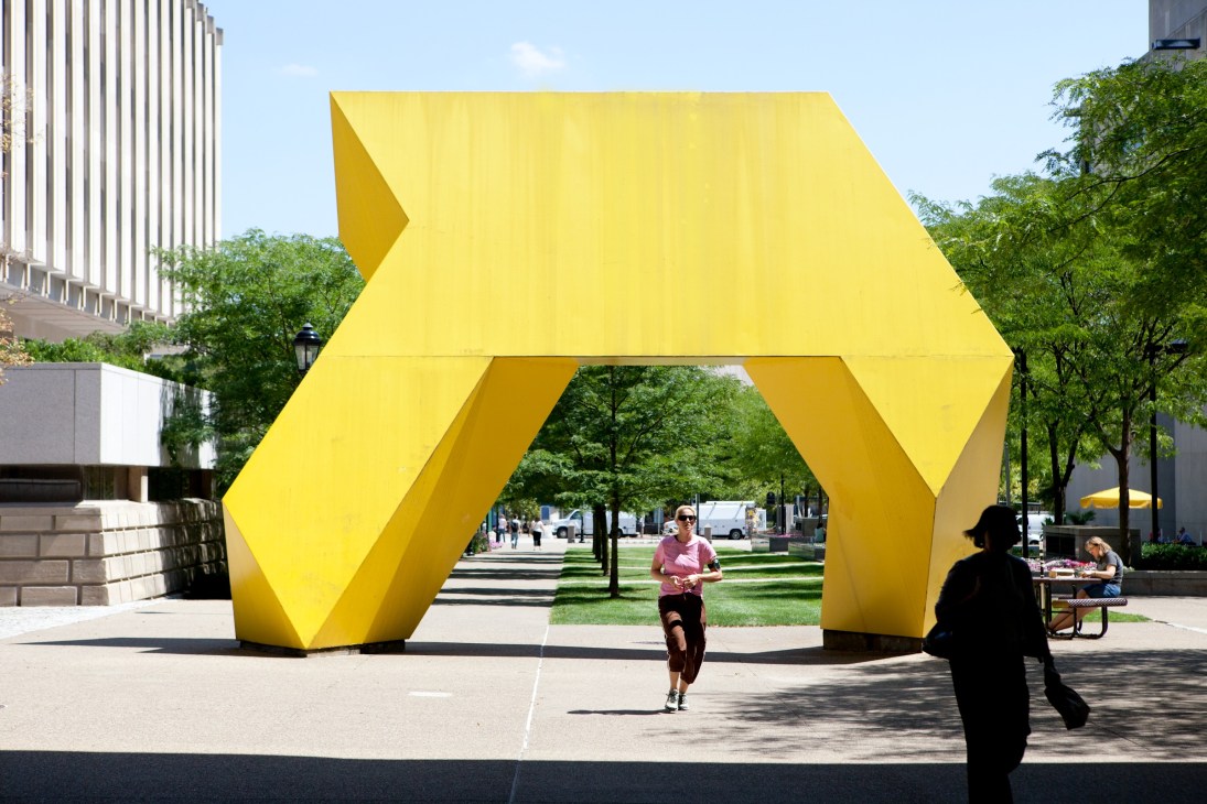 Yellow geometric arrow sculpture in an urban plaza with people walking beneath it.