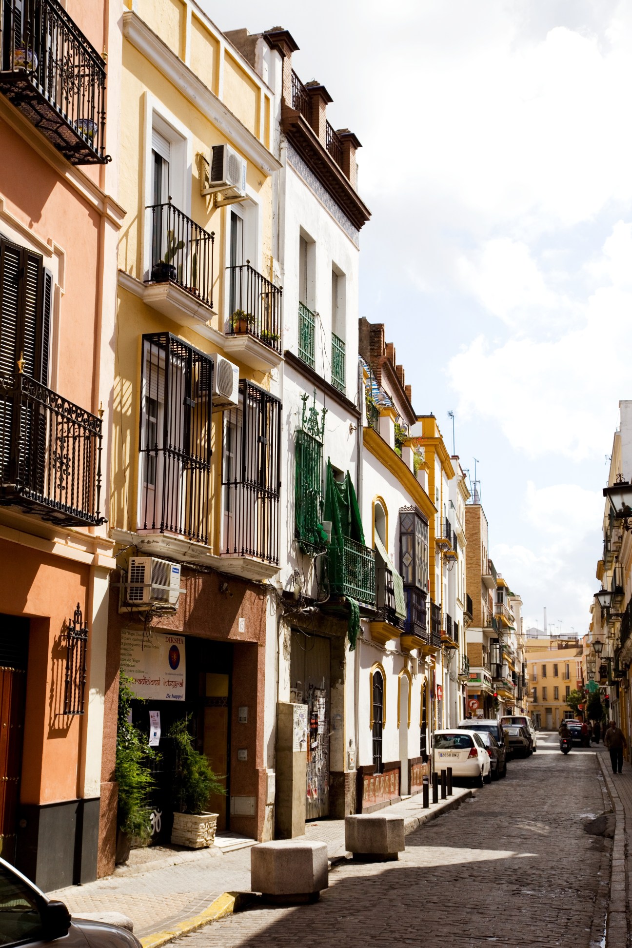 A sunny cobbled street in Seville lined with colorful historic buildings featuring wrought-iron balconies and wooden shutters.