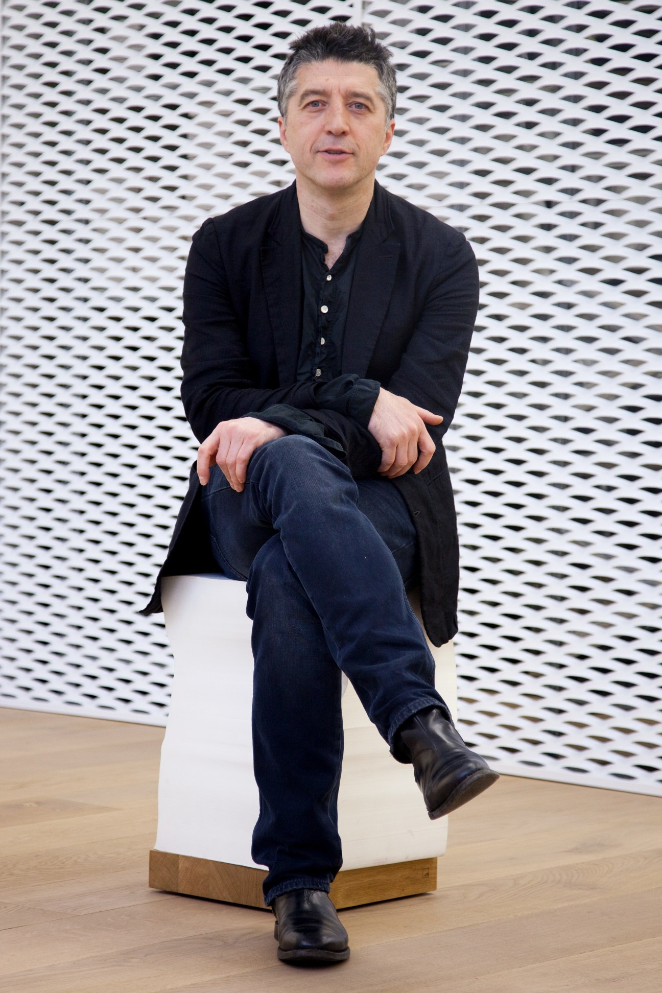 Andreas Murkudis sits on a white stool against a white perforated wall backdrop.