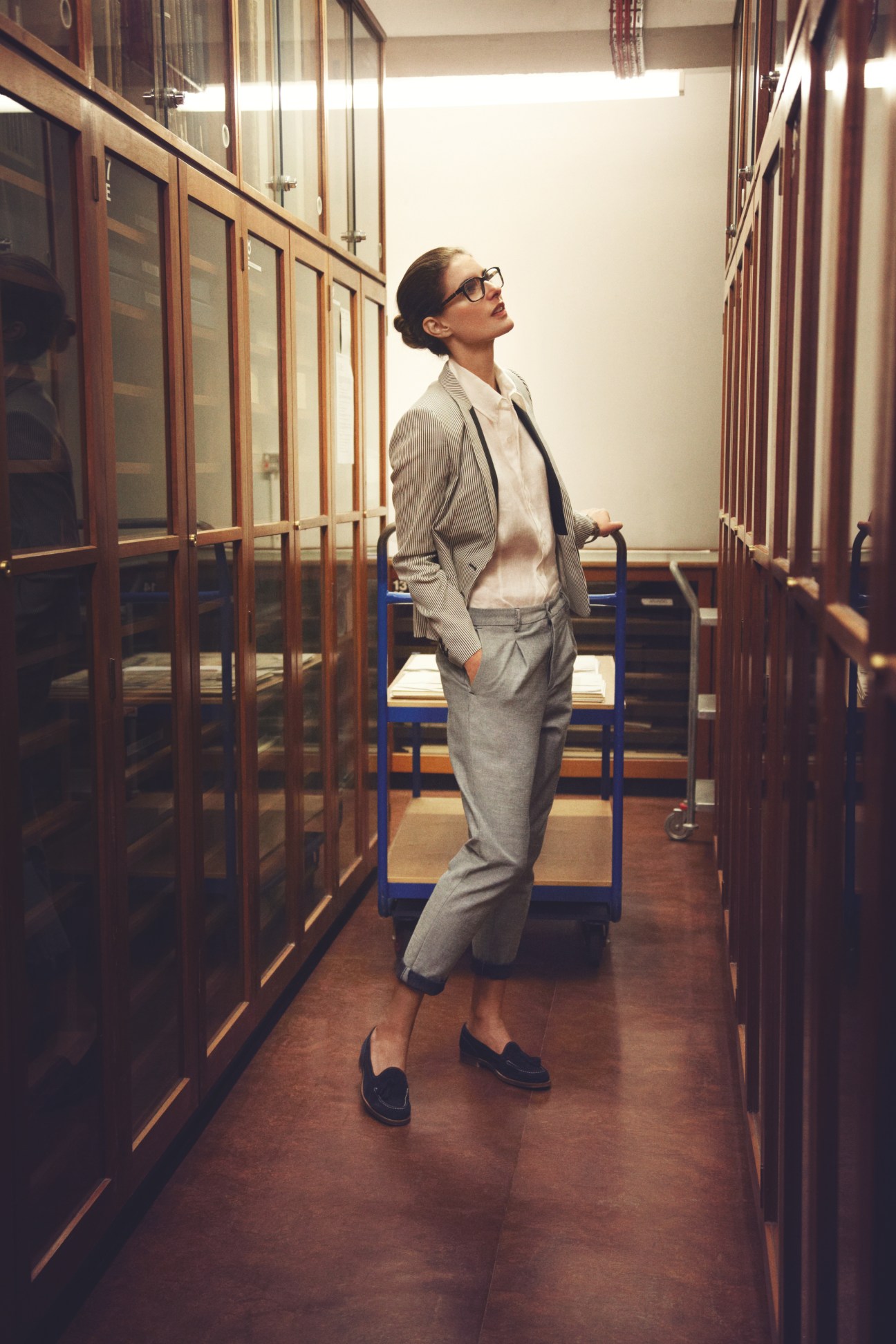 Woman in grey suit and Prada glasses standing in a library archive corridor between wooden cabinets.