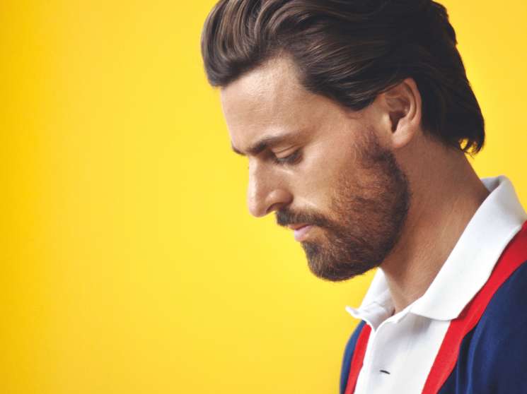 Man wearing Letroyes cardigan and Lacoste polo shirt against yellow background.