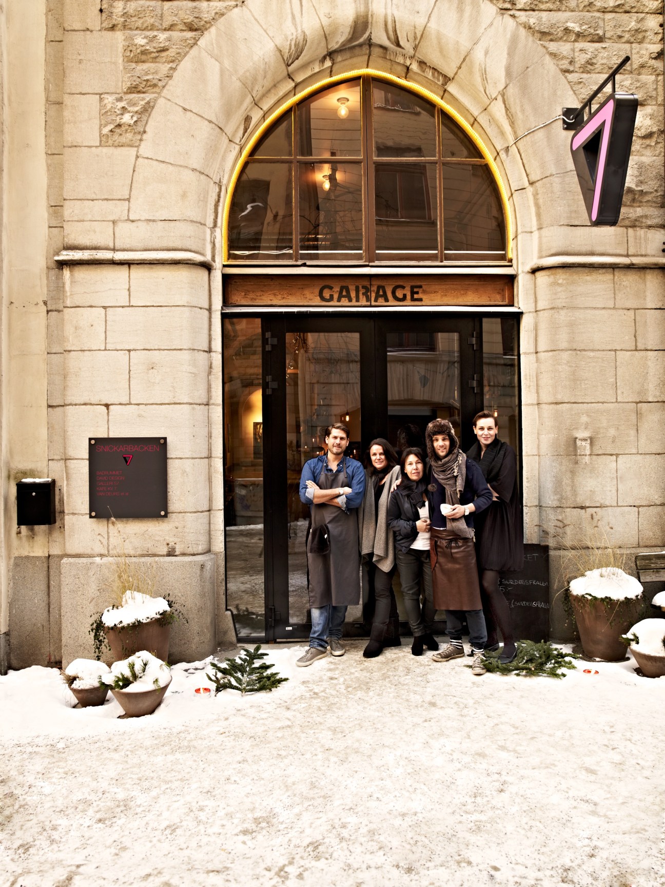 Five entrepreneurs stand in front of Snickarbacken 7's arched entrance with a yellow-lit window and "GARAGE" sign above.