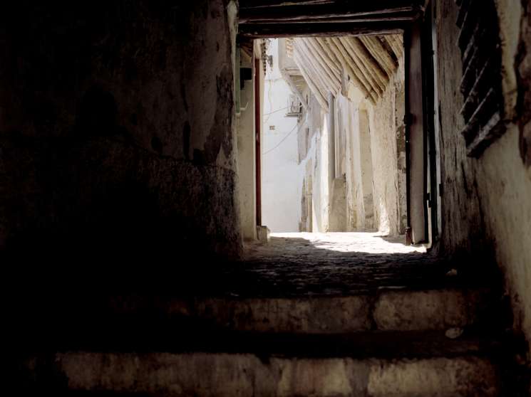 Dark narrow passageway opening onto sunlit street in Algiers casbah with weathered stone walls.