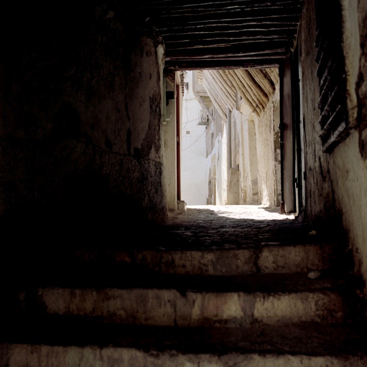 Dark narrow passageway opening onto sunlit street in Algiers casbah with weathered stone walls.