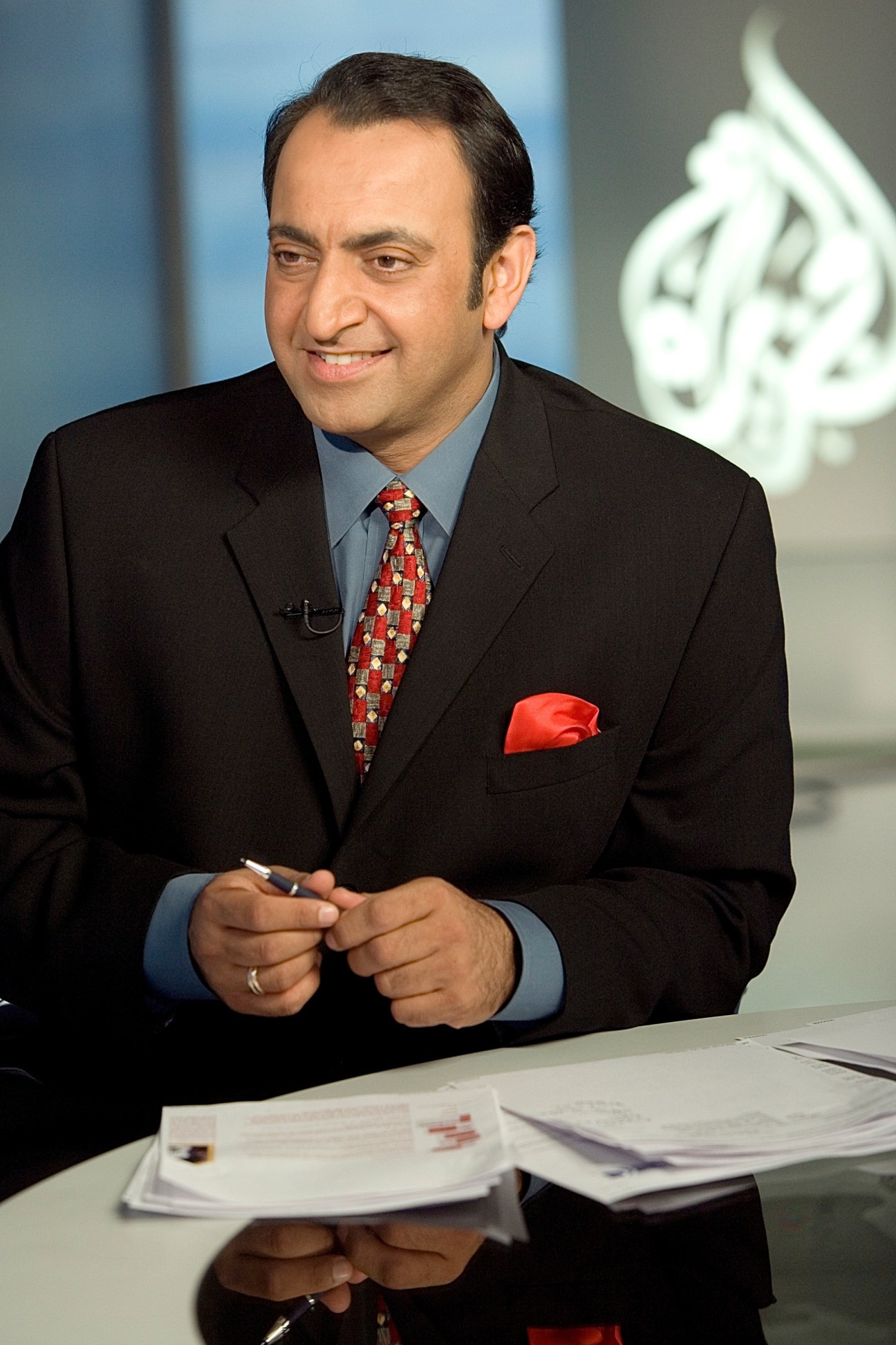 Man in business suit and red tie at news desk with Al Jazeera logo visible in background.