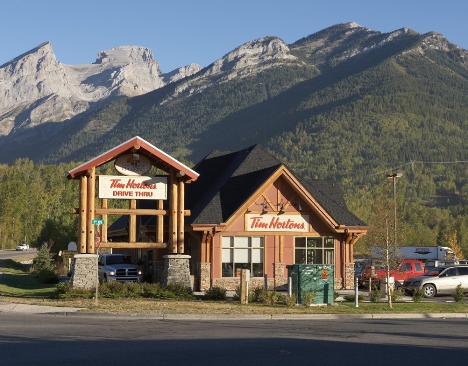 Tim Hortons drive-thru building with mountain backdrop and parked vehicles.