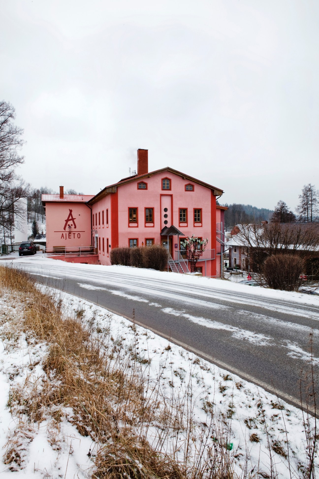 Pink-painted Ajeto glassmaking factory building in snowy landscape with forested hills in background.