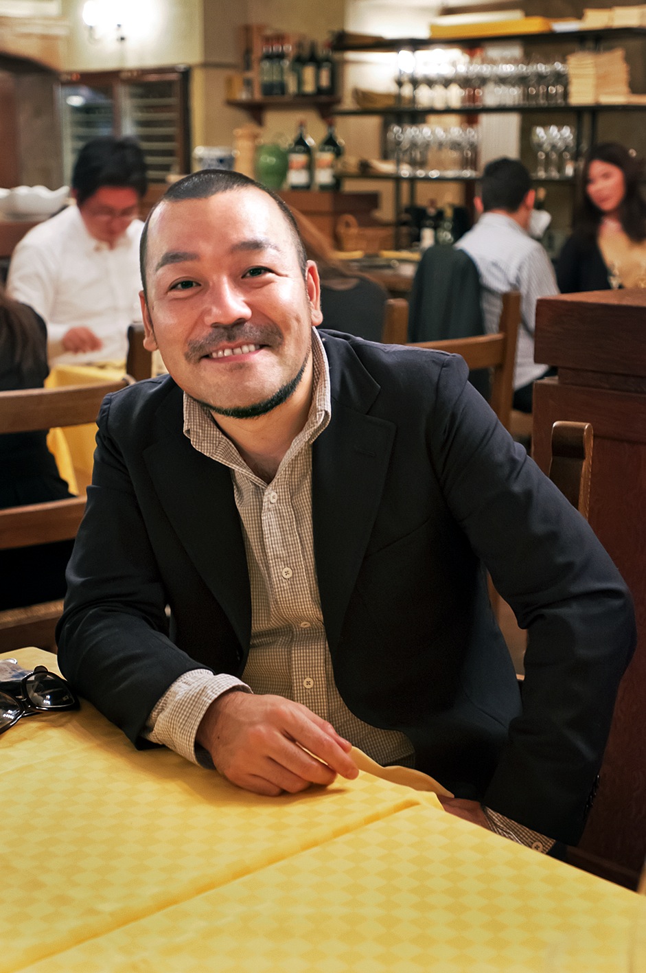 Kentaro Kobayashi smiling at a table inside La Bisboccia restaurant with diners and shelved bottles visible in the background.