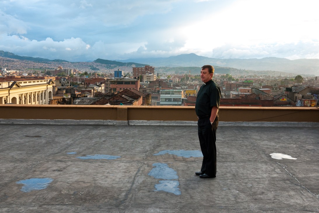 Priest Dario Echeverri standing on a rooftop overlooking the sprawling cityscape of Bogotá, Colombia, with mountains in the distance.