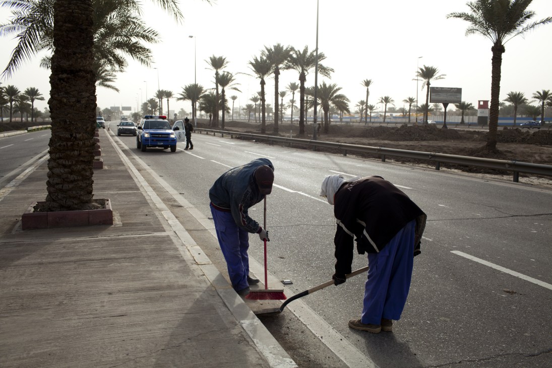 Workers sweep the Baghdad Airport Road lined with palm trees and traffic barriers.