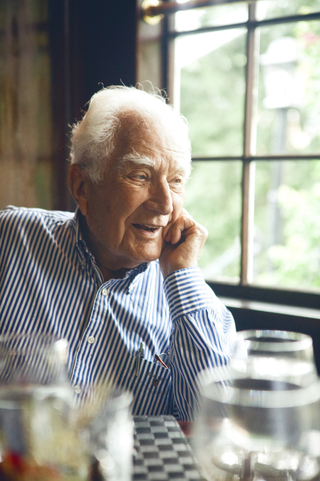 Elderly man with white hair in blue striped shirt sitting at a dining table by a window.