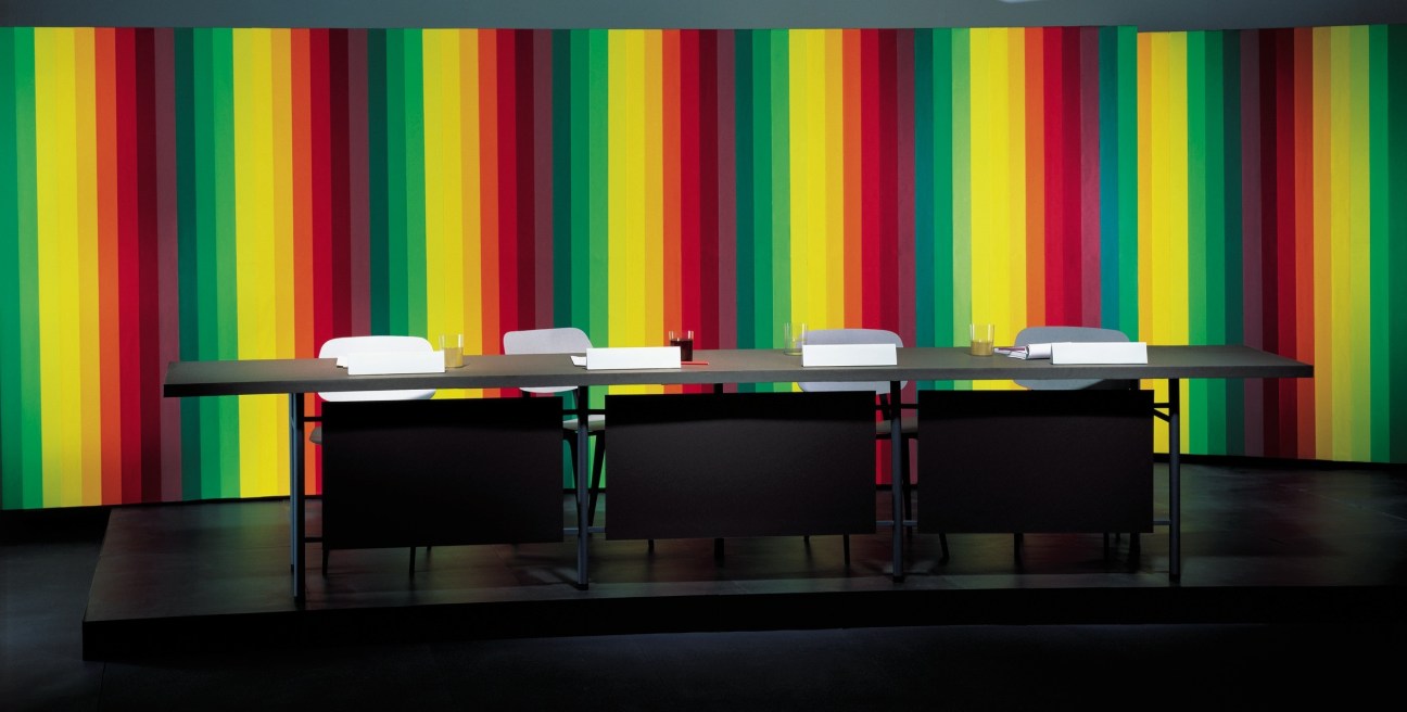 Modern dining table with black chairs against a colorful striped wall installation by Thomas Demand.