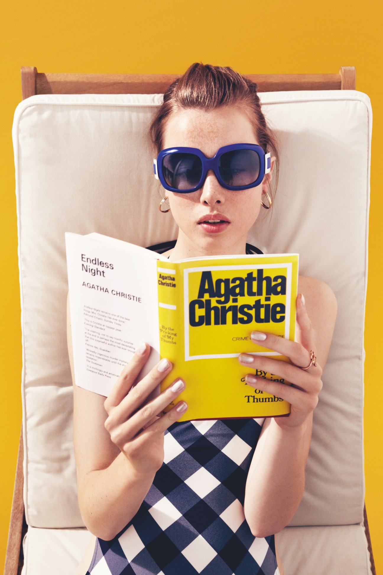Model wearing blue Roger Vivier sunglasses and Cartier earrings, reading Agatha Christie book on lounger