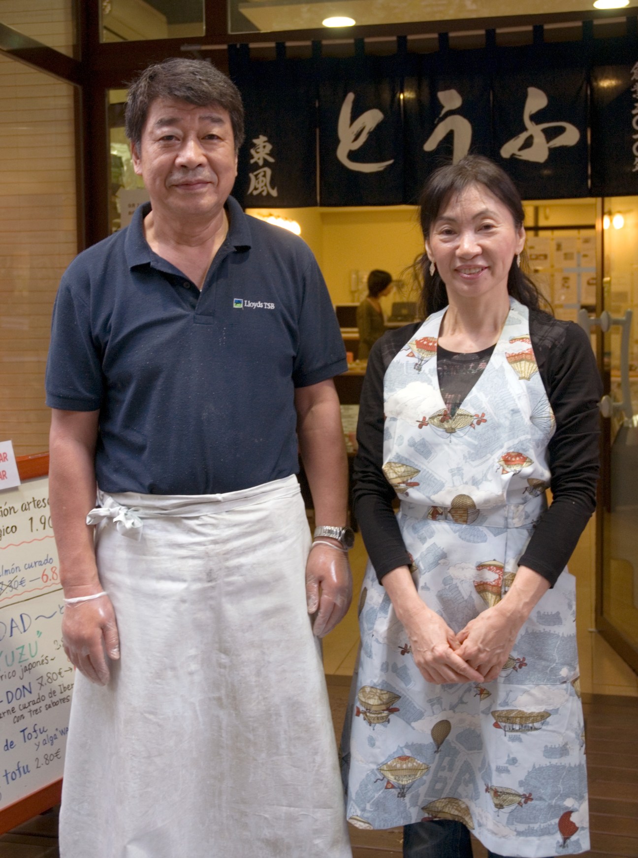 Tateo and Michiko Shimizu stand together in their tofu shop in Barcelona, with Japanese signage visible behind them.