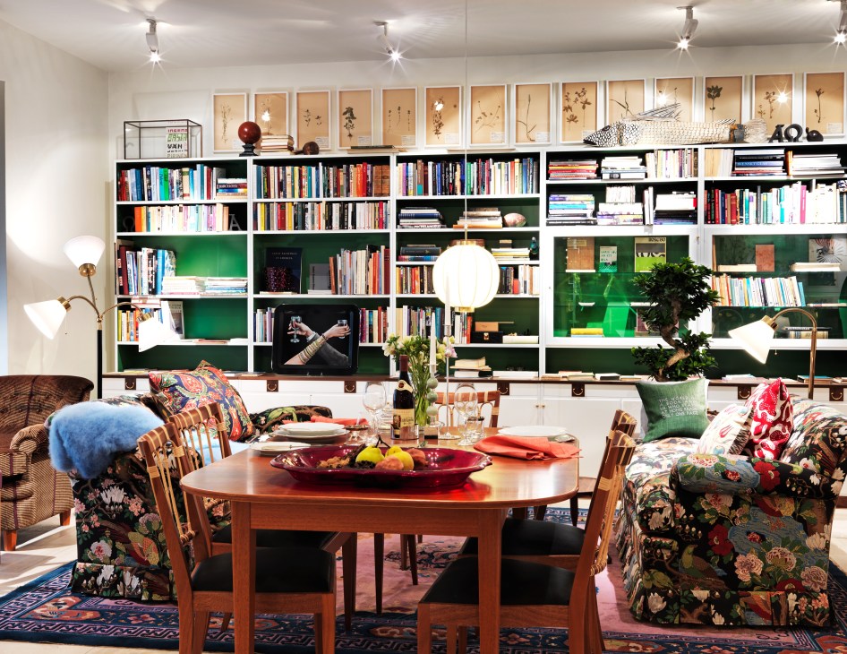 Living room display at Svenskt Tenn showing round wooden table, patterned upholstered chairs, colorful textiles, and white shelving with books and decorative objects.