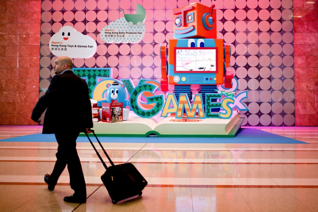 A man with luggage walks past a large colorful robot mascot display at the Hong Kong Toys & Games Fair entrance.