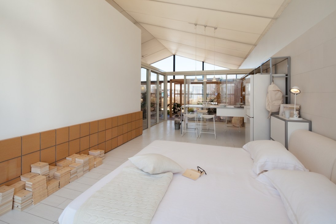 Modern bedroom with white bed, orange wall paneling, and glass panels overlooking an open-plan living area with skylights.