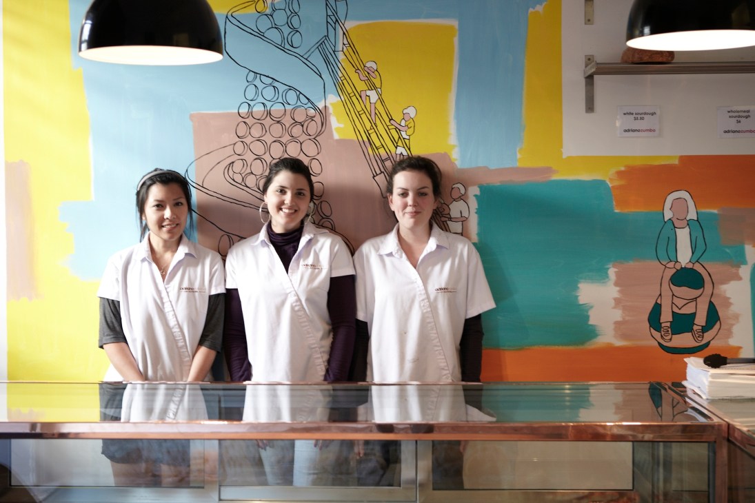 Three staff members in white uniforms standing behind a counter at Adriano Zumbo patisserie in Sydney.
