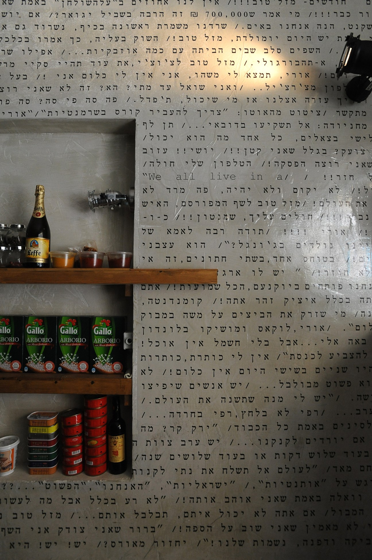 Interior of Machneyuda restaurant with Hebrew text on wall, wooden shelves displaying bottles and canned goods.