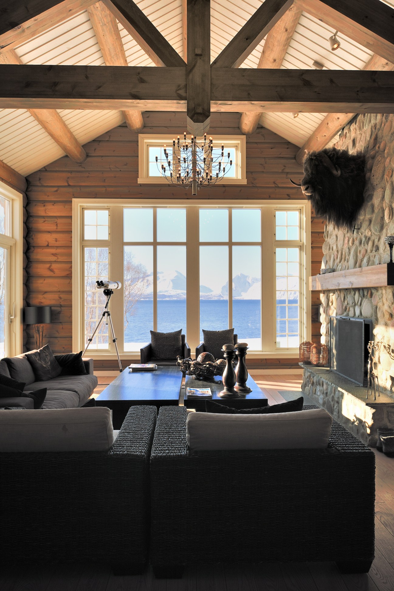 Interior of Lyngen Lodge with wooden beams, large windows overlooking a fjord and mountains, black furniture, and stone fireplace.