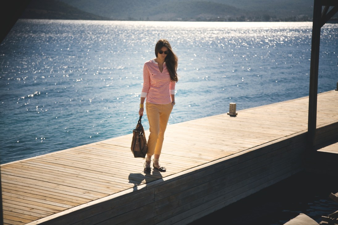 Woman in striped t-shirt and trousers standing on wooden deck overlooking sparkling Mediterranean sea.