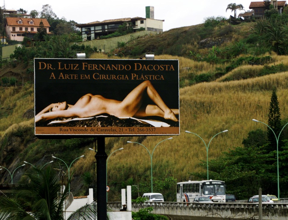 Roadside billboard advertising plastic surgery services featuring an airbrushed female figure in Brazil.
