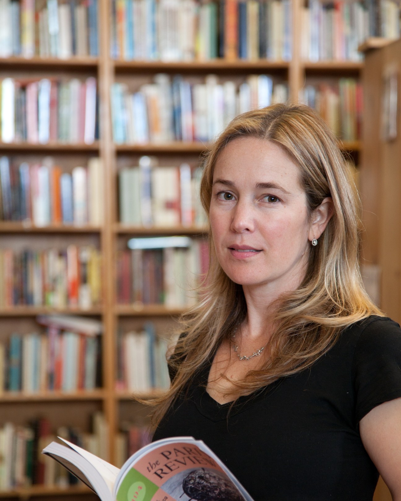 Author Vendela Vida holds The Paris Review magazine while standing in front of bookshelves.