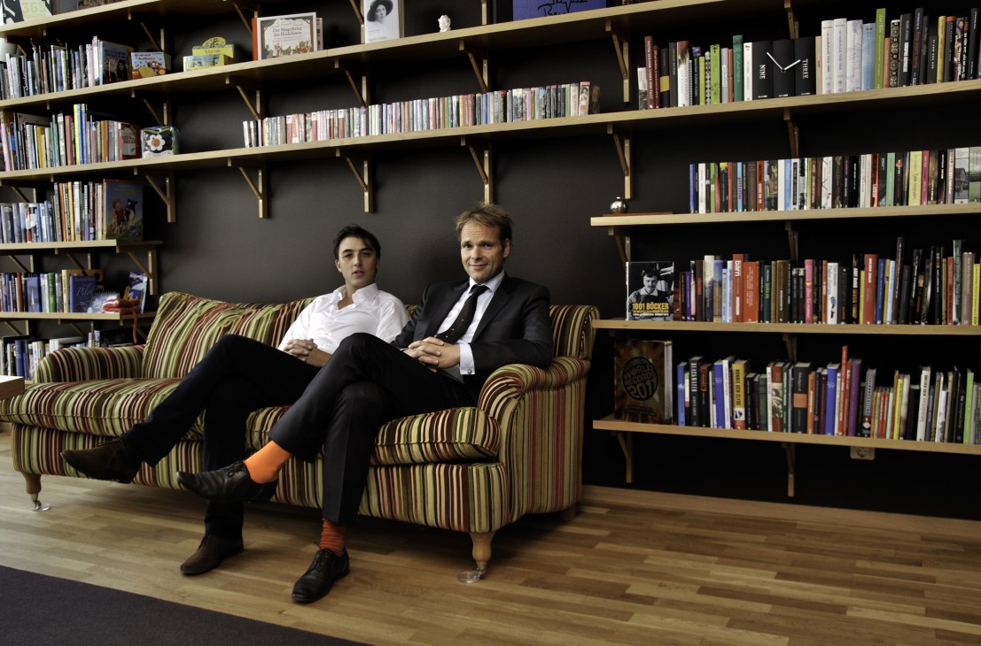Two men sit on a striped sofa against a wall of bookshelves in an office setting.