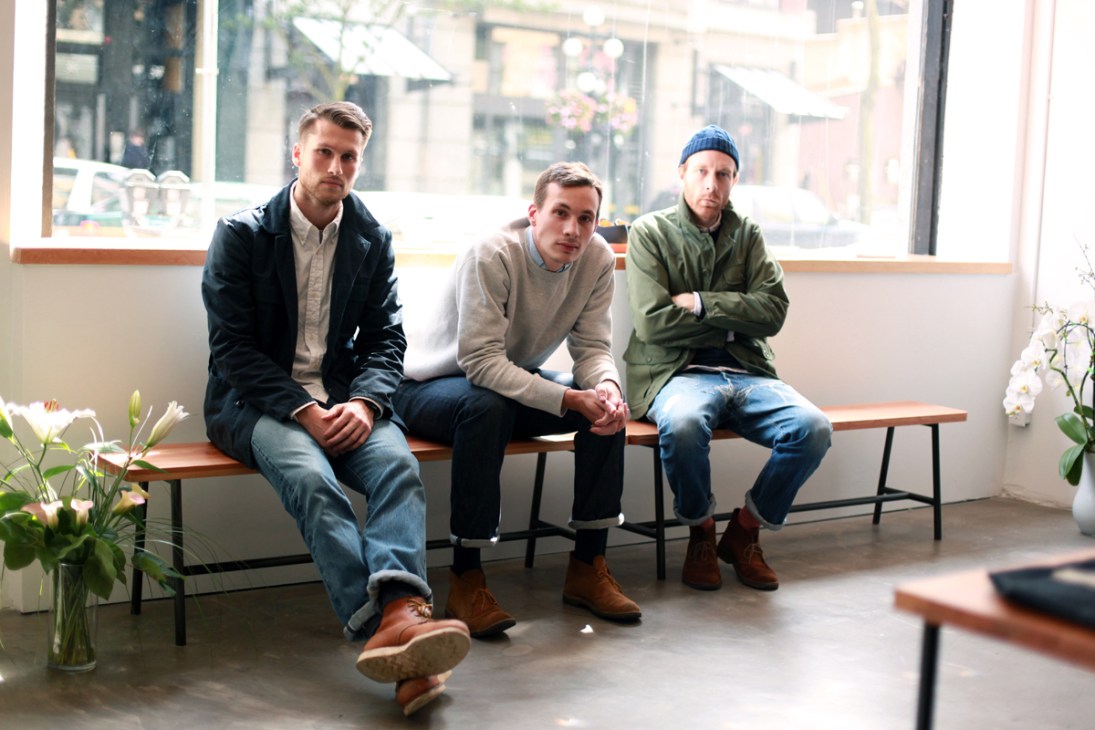 Three men sit on a wooden bench by a large window in a minimalist retail space.
