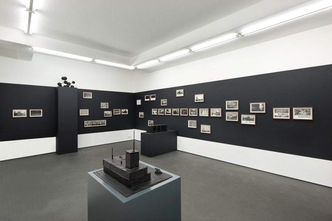 Gallery interior with black and white walls displaying framed photographs and a sculptural object on a pedestal.
