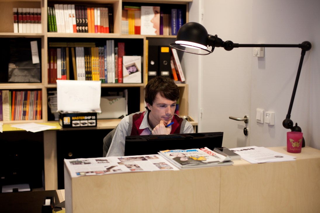 Man working at a desk with magazine layouts, shelves of colorful books behind him and a black desk lamp overhead.