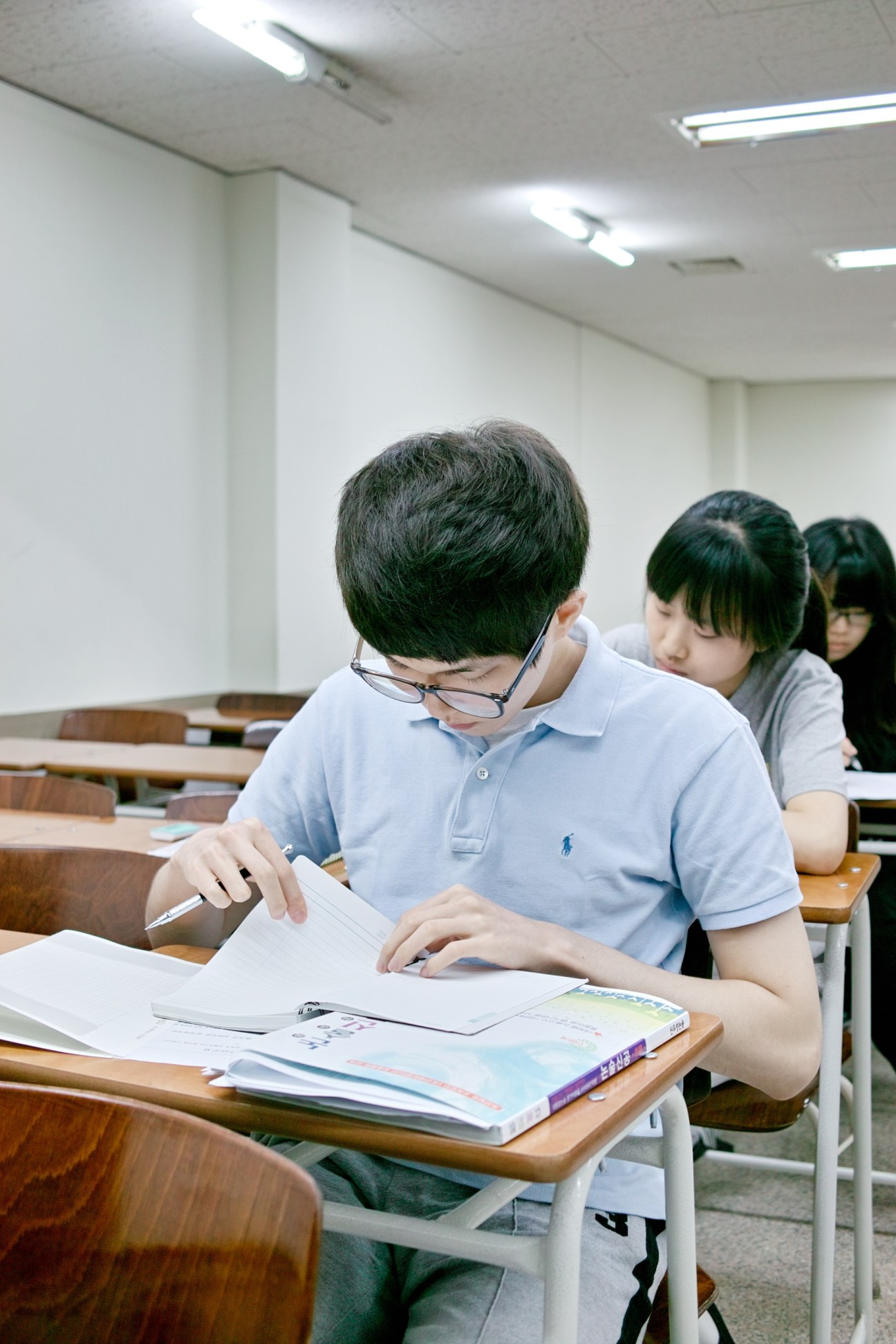 Young student in glasses wearing light blue shirt reviews test paper in Korean language classroom.