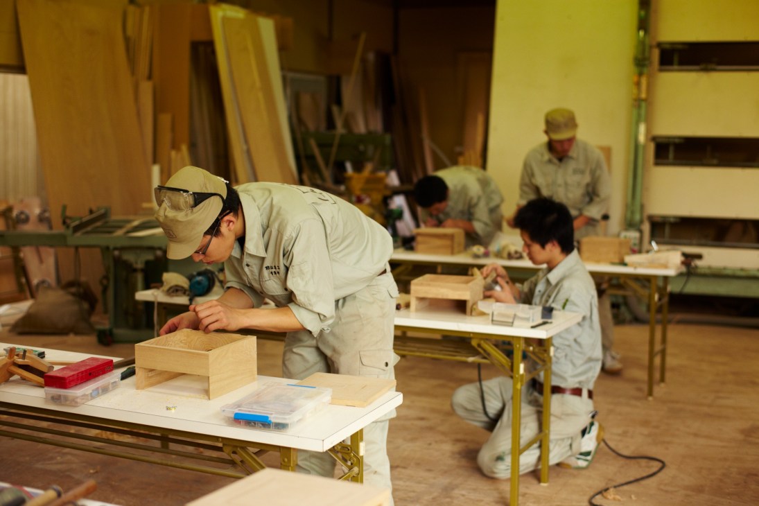Woodworking apprentices in beige uniforms practice furniture-making techniques at workbenches in a workshop.
