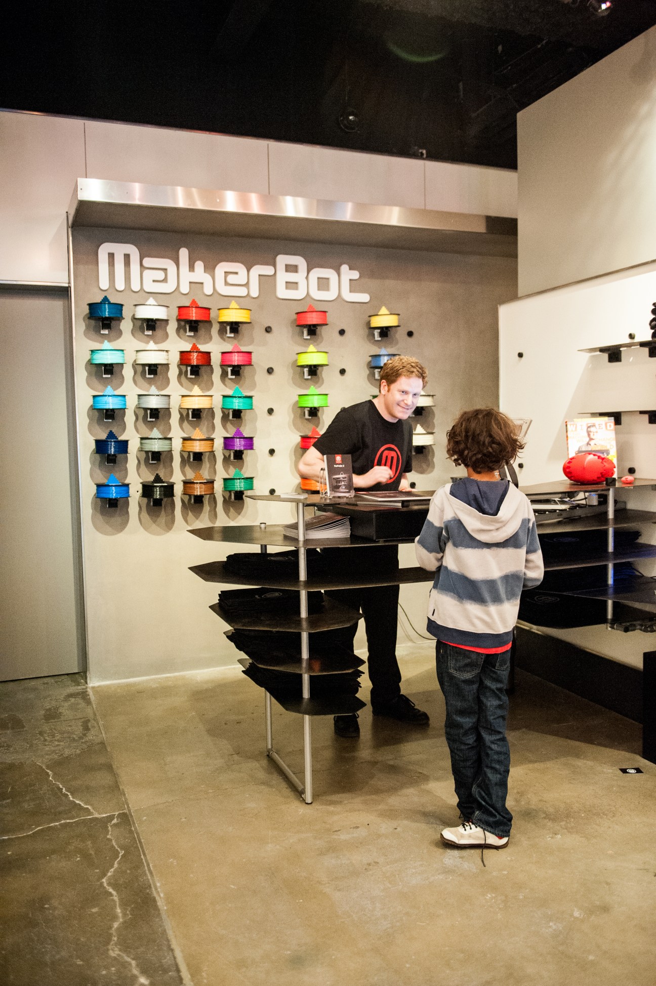 MakerBot retail store interior with colorful 3D printer heads displayed on wall and staff assisting customer.