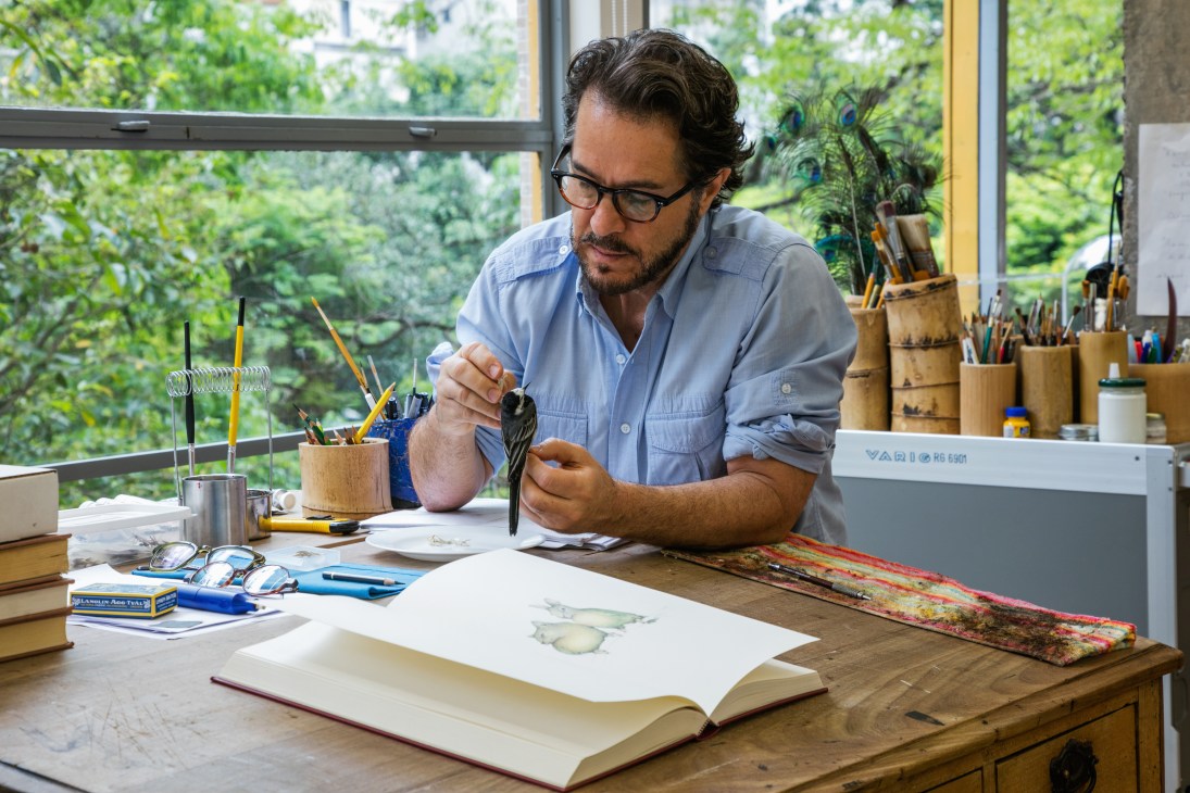 Artist Walmor Corrêa working at his desk in a bright studio space with large windows overlooking greenery.