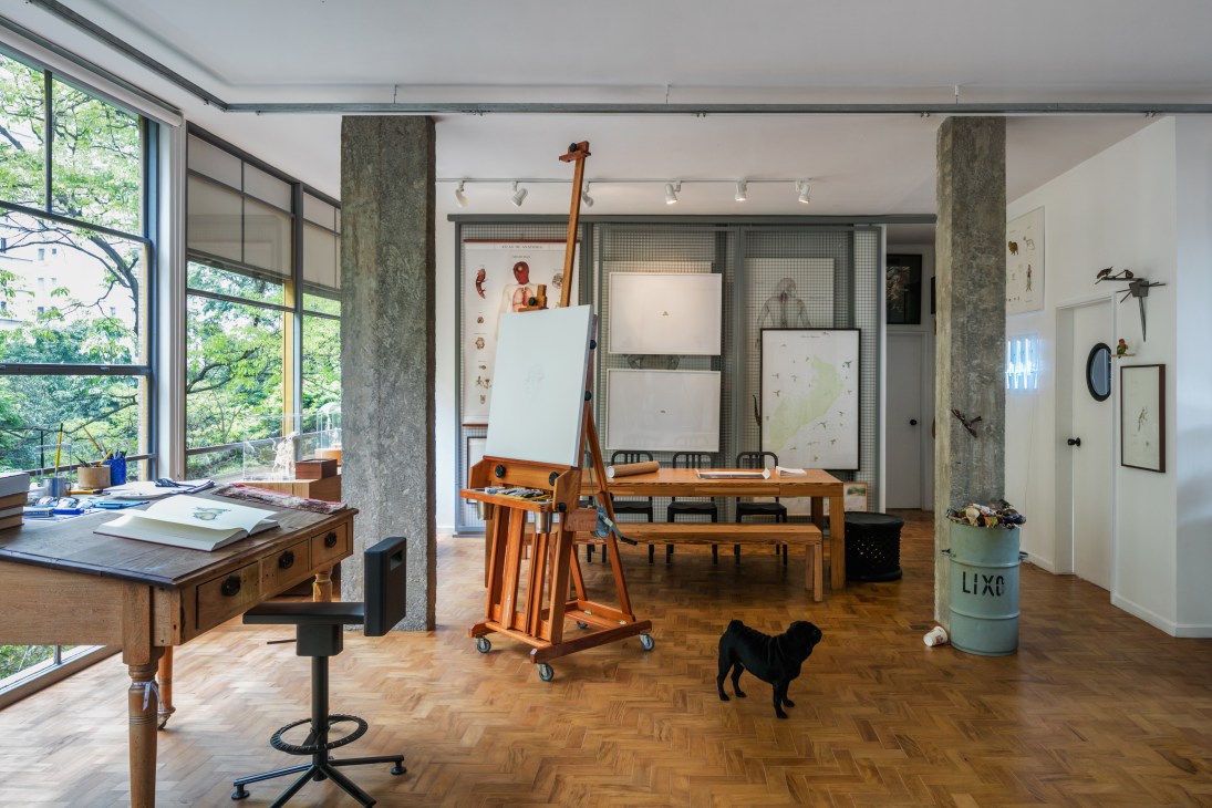 Artist Walmor Corrêa's living room and atelier with wooden furniture, large windows overlooking greenery, and a black pug on parquet flooring.
