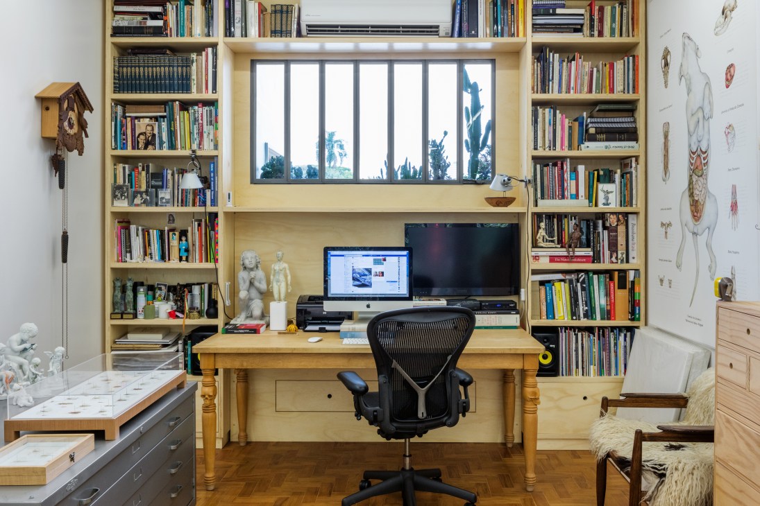 Office with wooden desk, built-in shelving filled with books, computer setup, and succulents on the window sill overlooking greenery.