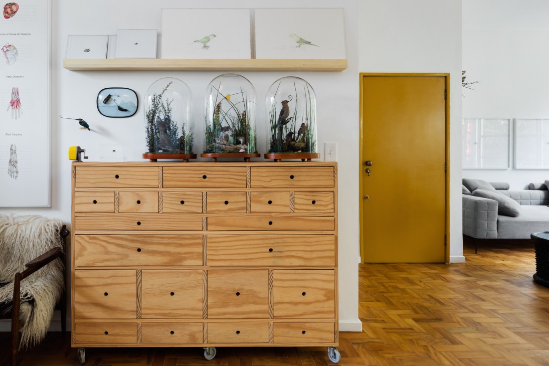 Wooden storage drawers with wheels holding glass terrariums and artwork beneath a wooden shelf and mustard-yellow door.