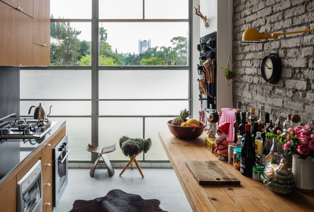 Modern kitchen with exposed brick wall, wooden counters, gas stove, and large windows overlooking São Paulo's green landscape.