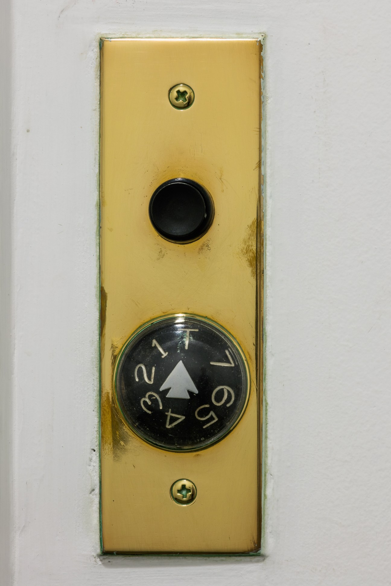Vintage yellow lift call button plate with black dial marked with floor numbers and white directional arrow.