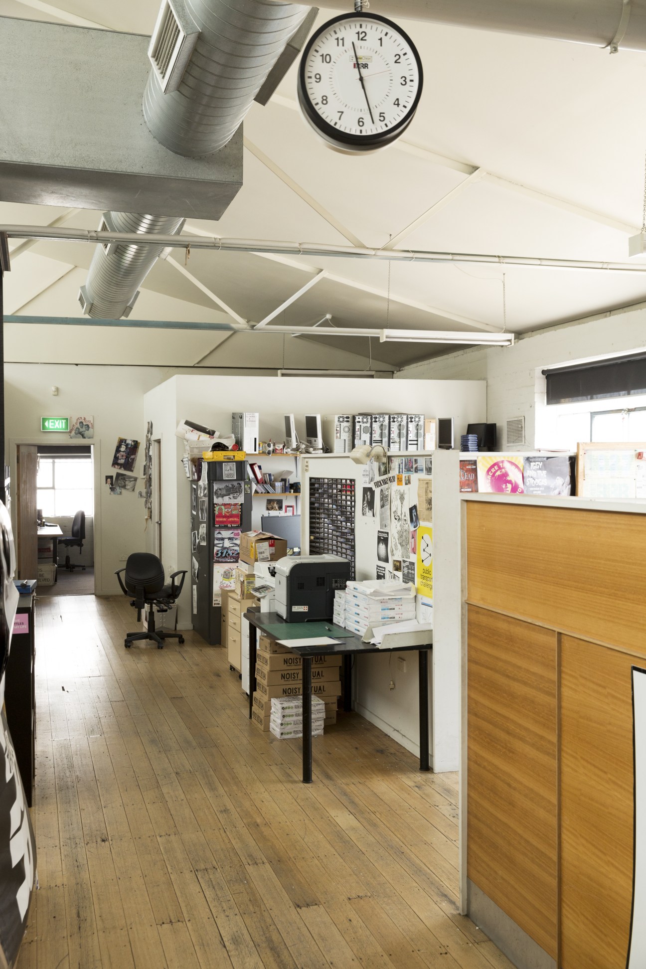 Interior view of Triple R radio station in Brunswick showing office space with clock, desk, shelving and promotional materials.