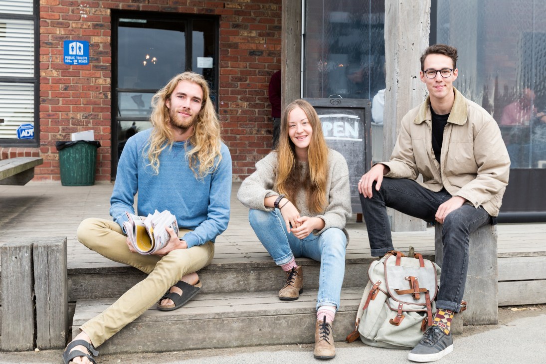 Three young people sitting outside Wye River General Store on wooden steps, holding magazines and a backpack.