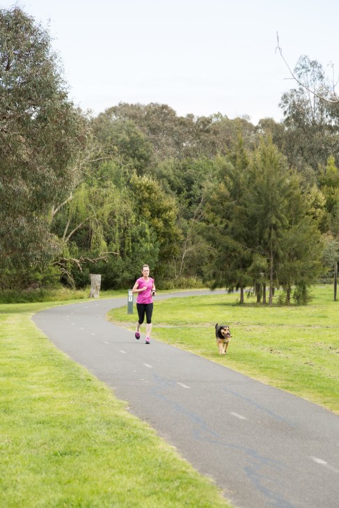 Woman jogging with a dog on a paved path through parkland with trees and green grass.