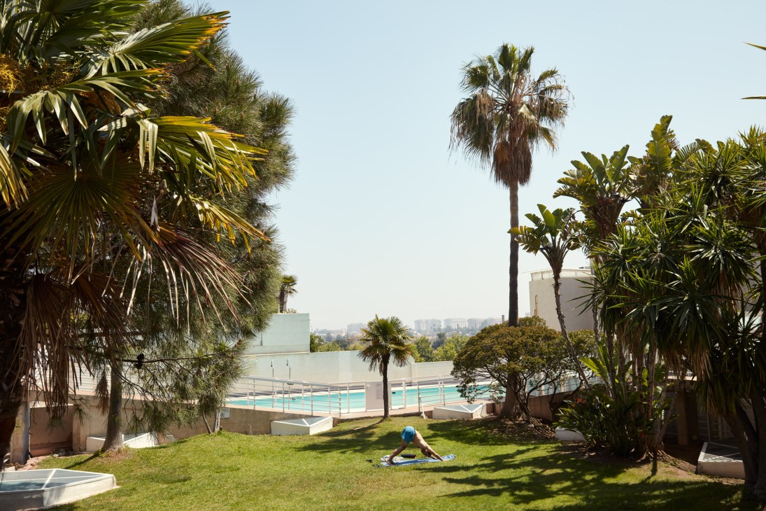 Person doing yoga on a grassy tropical garden terrace with palm trees and pool visible behind brutalist building.